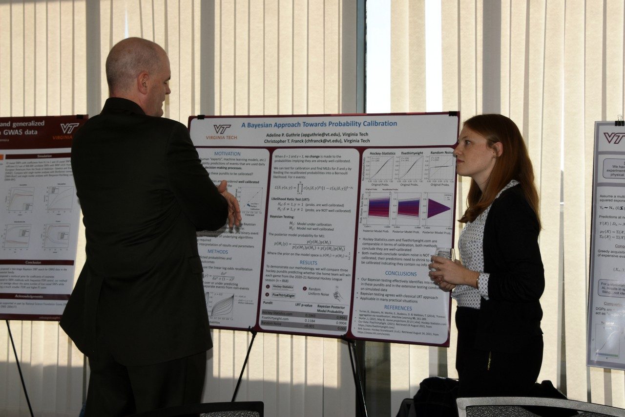 Two individuals in conversation stand in front of a project poster.